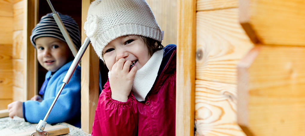 Zwei Kinder mit Wintermützen lachen in einem Holz-Spielhaus